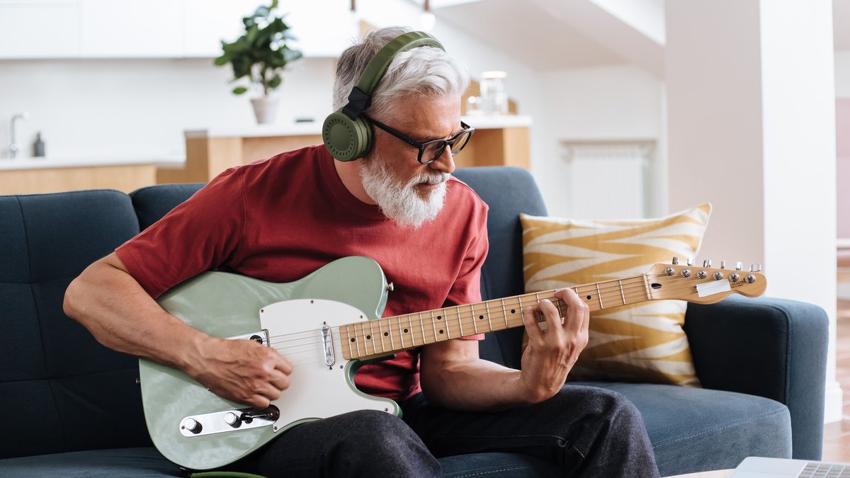 Elderly man enjoys playing electric guitar at home, using headphones for a personal music session.