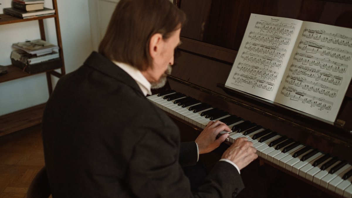 Senior man in formal attire playing piano indoors, focused on sheet music.