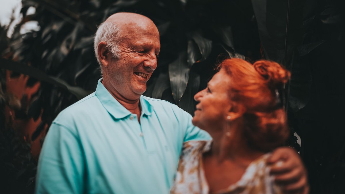 Elderly couple smiling and embracing in lush outdoor setting, capturing love and friendship.