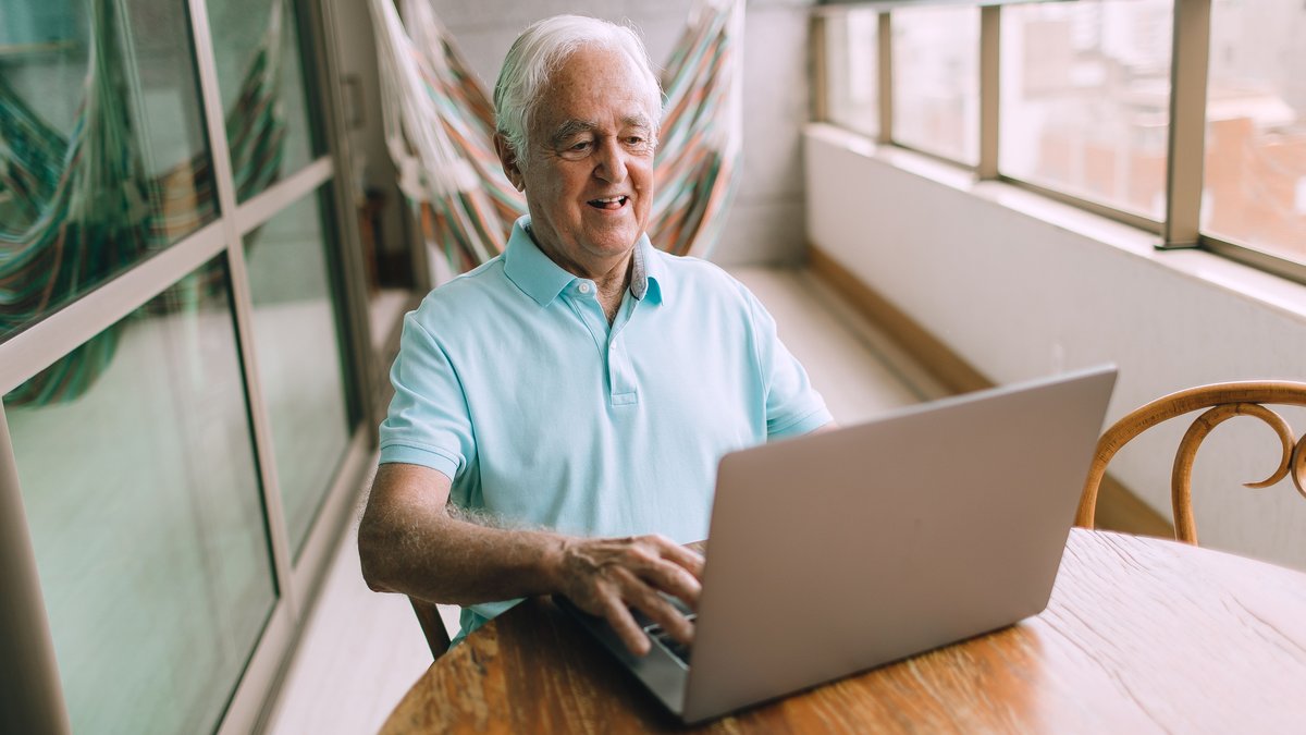 Elderly man happily using a laptop on a sunny balcony, embracing online connectivity.