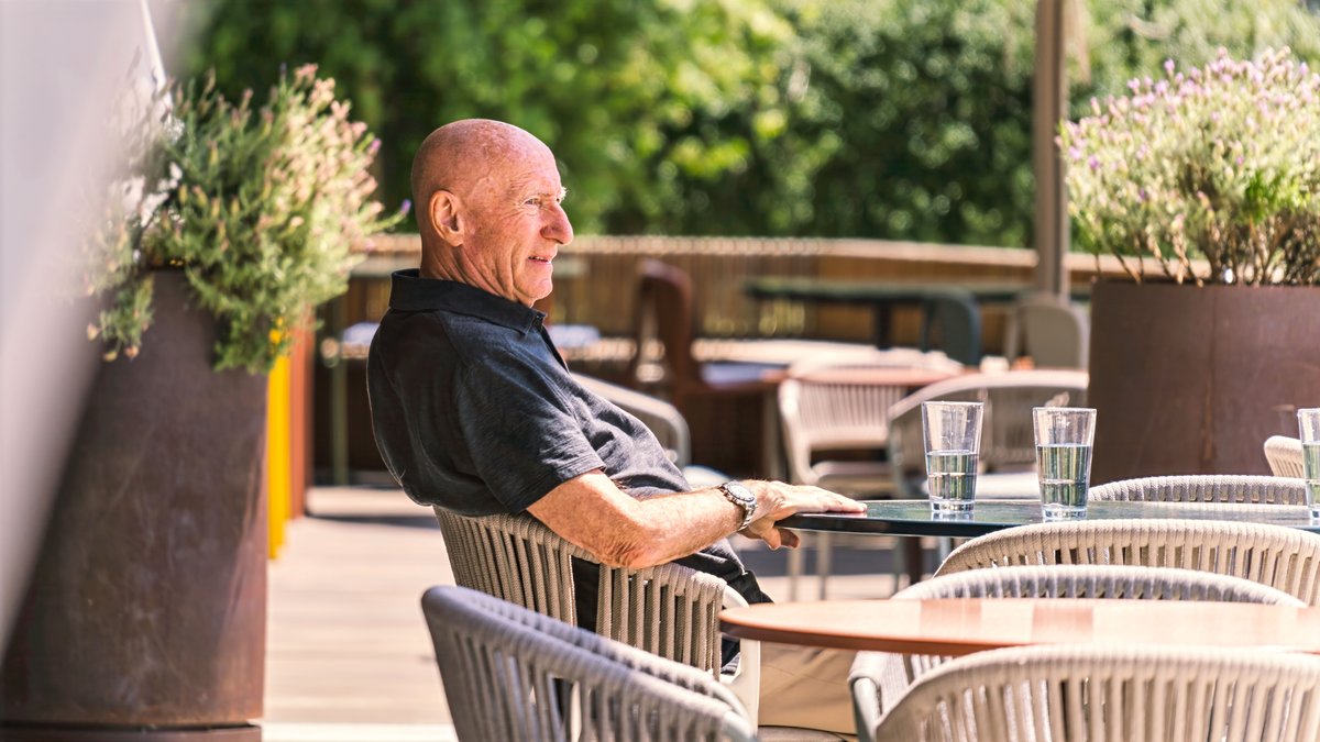Senior man sitting at an outdoor patio, enjoying a sunny day with drinks on the table.