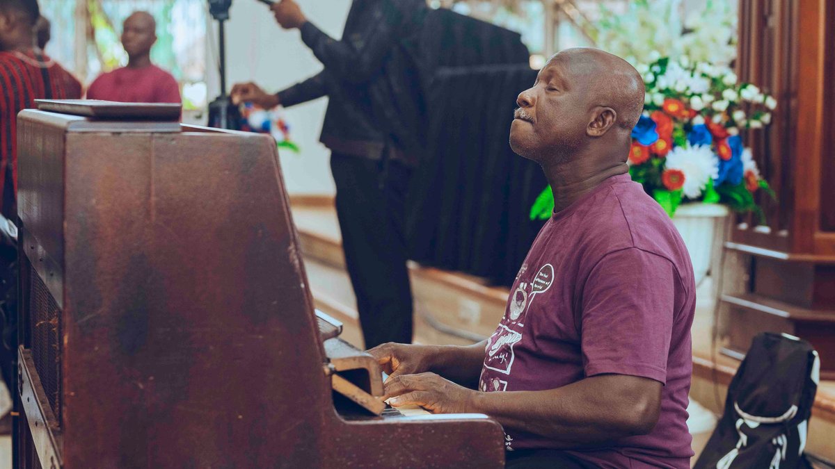An elderly man playing a piano at an indoor event filled with people and vibrant floral decorations.