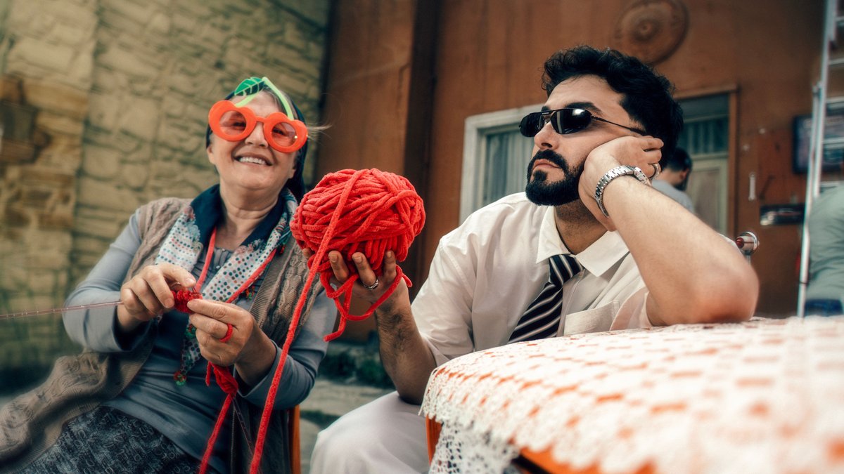 A cheerful senior woman knitting with a young man holding yarn, capturing a joyful outdoor moment.