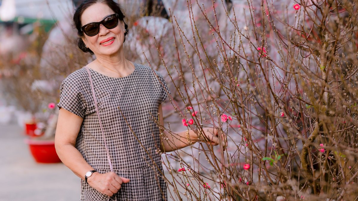 Elegant senior woman smiles amidst blooming cherry blossoms in a sunny garden.