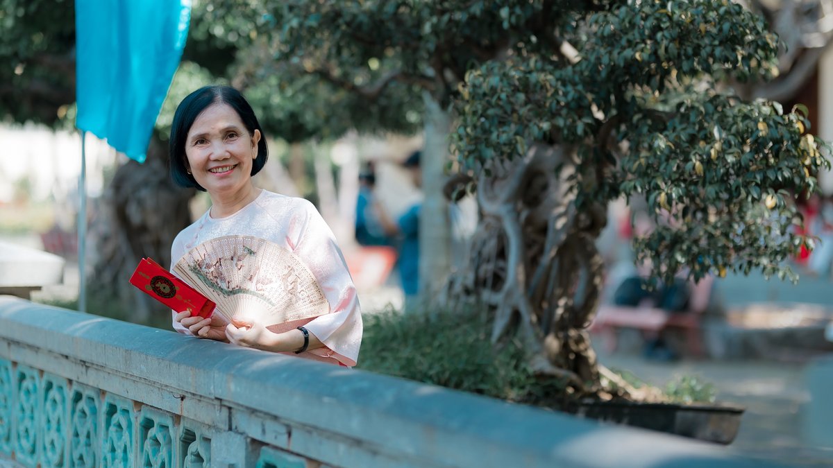 Smiling elderly woman holding a fan and red envelope outdoors in a sunny park.