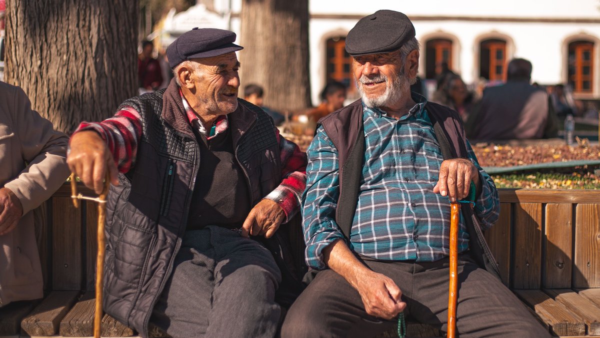 Two senior men wearing hats sit on a bench, chatting and laughing on a sunny day.