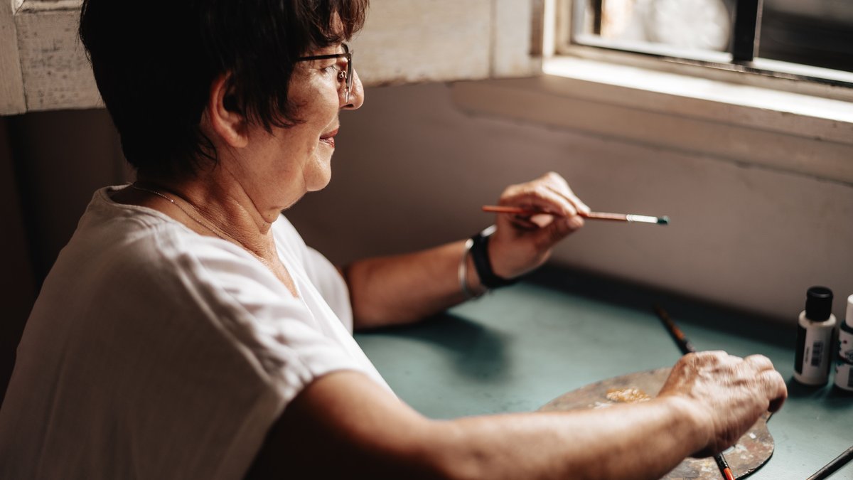 A senior woman enjoys painting near a window, illustrating creativity and leisure.