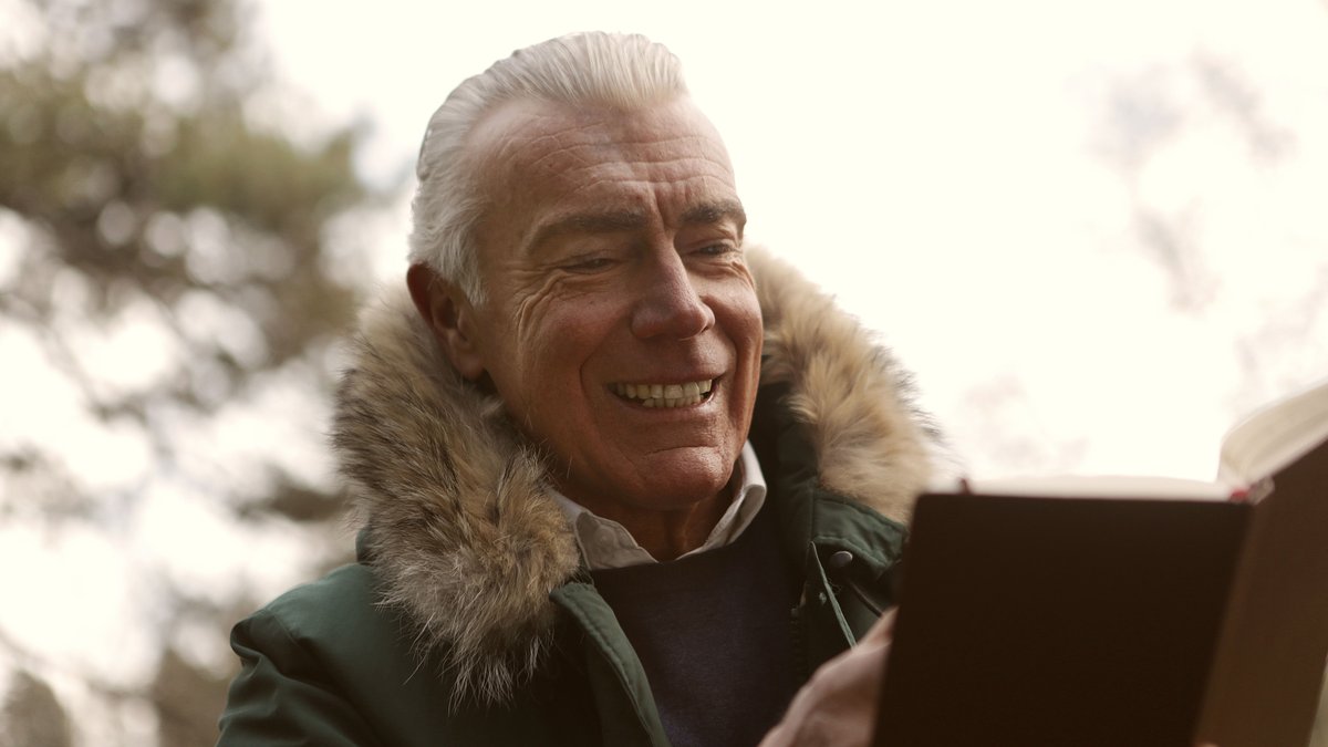 A cheerful senior man in a fur-lined coat enjoys reading a book outdoors, capturing leisure and joy.