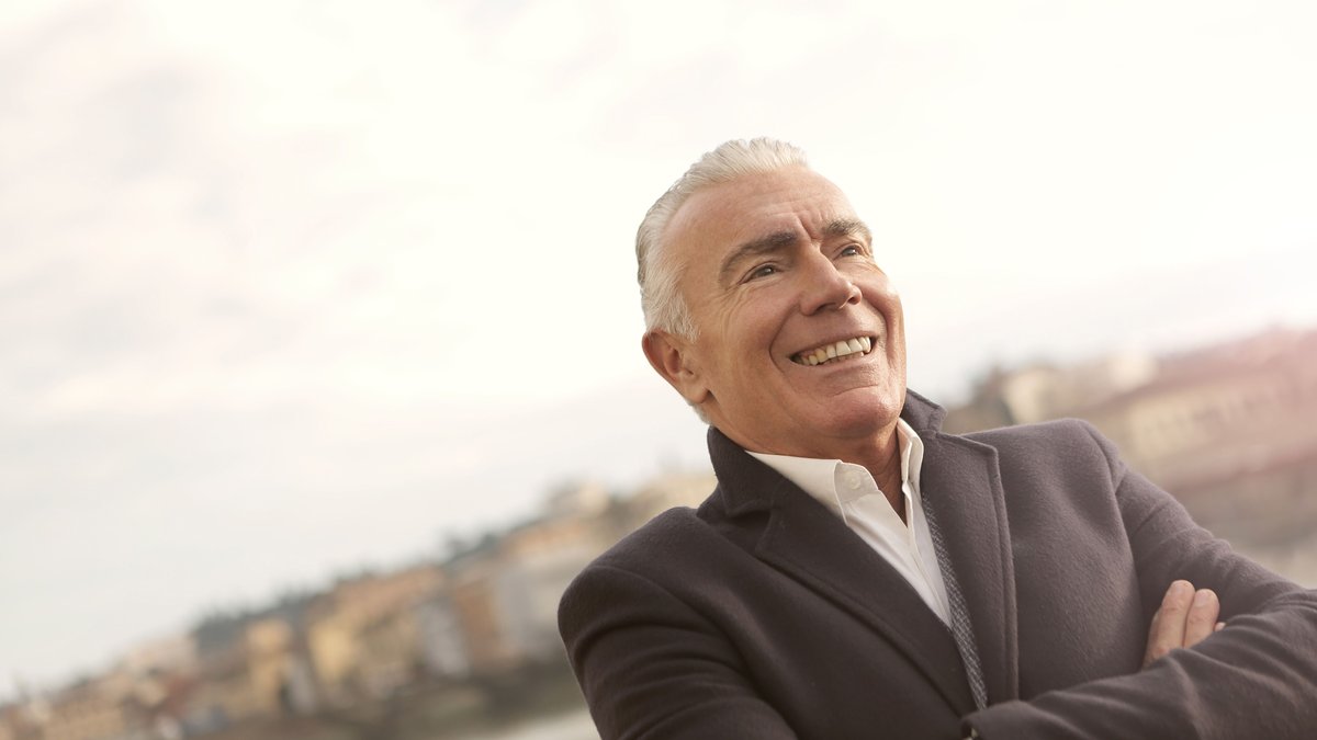 A senior man in business attire smiles confidently during a sunny outdoor setting.