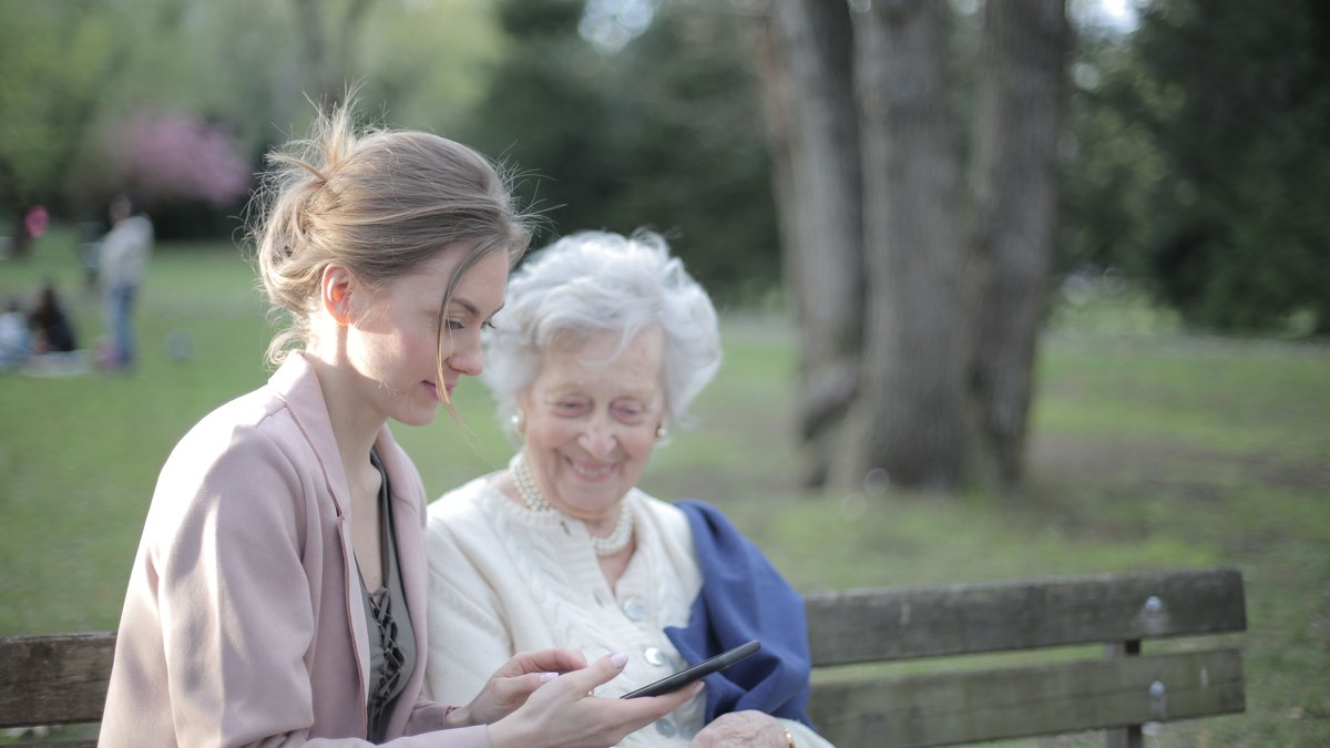 Side view of smiling adult female helping aged mom in using of mobile phone while sitting together in park