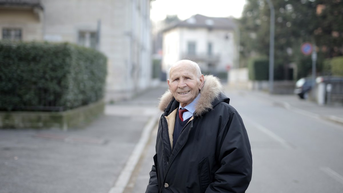 A cheerful elderly man in a coat smiling while standing outdoors on a city street.