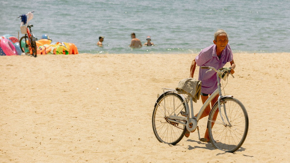 An elderly man pushes a bicycle along a sunny, sandy beach with people swimming in the background.