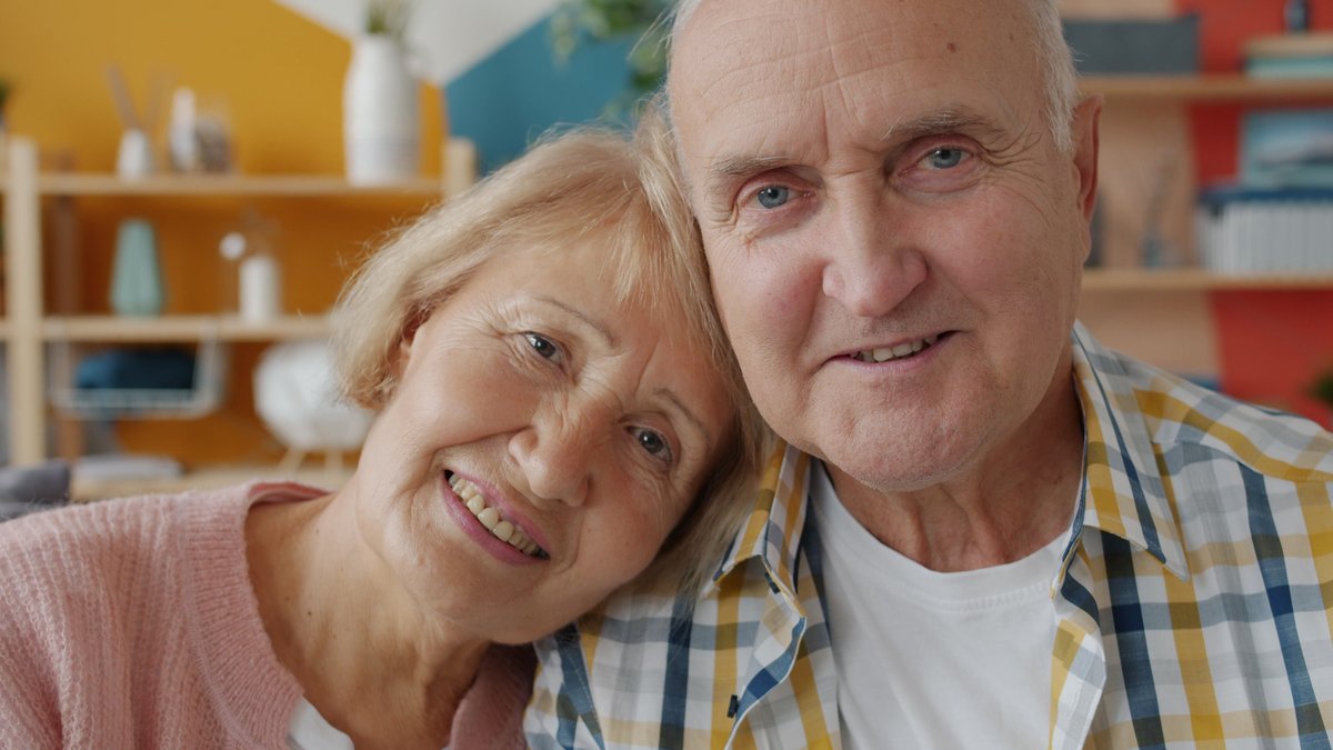 Charming elderly couple smiling warmly while seated indoors, enjoying a cozy moment together.