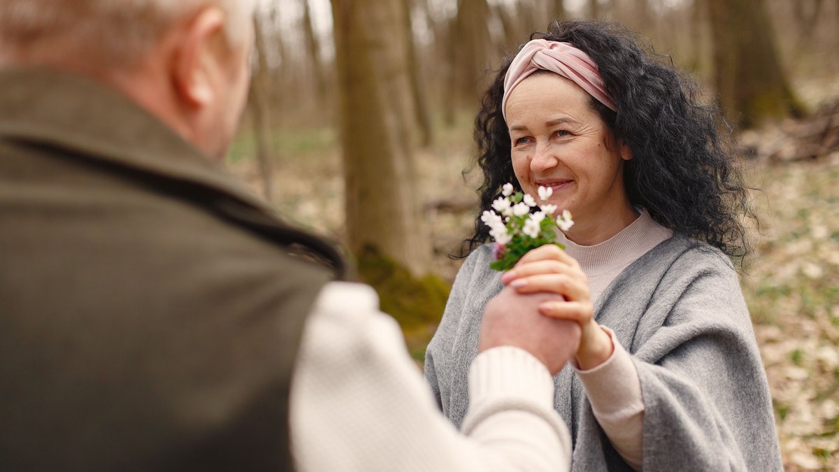 Crop elderly gray haired husband in warm clothes presenting bunch of fresh flowers to cheerful senior wife wearing wide scarf and headband while spending time together in autumnal forest