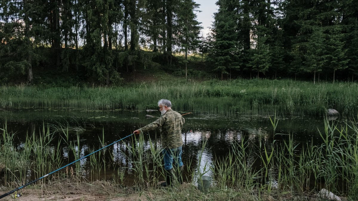 An elderly man enjoys a peaceful fishing session at a serene riverbank surrounded by lush greenery.