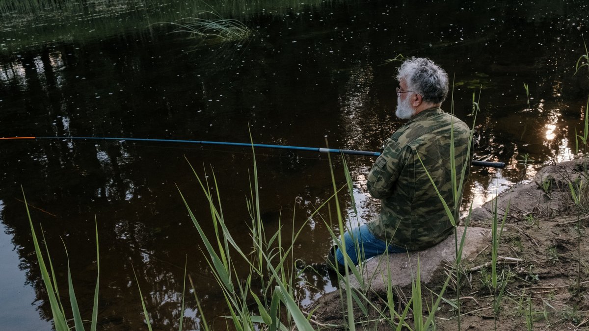 Senior man in camo jacket enjoys quiet fishing at a serene riverbank surrounded by nature.