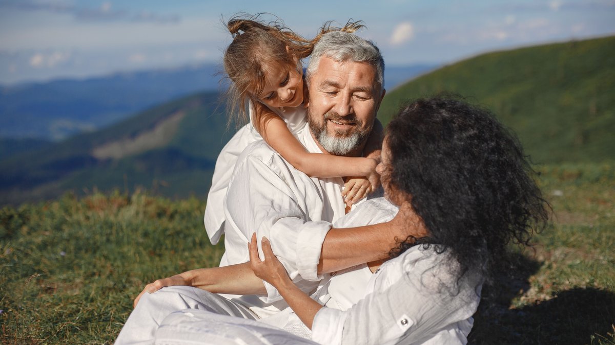 A joyful family moment outdoors, showing grandparents hugging their granddaughter under a sunny summer sky.