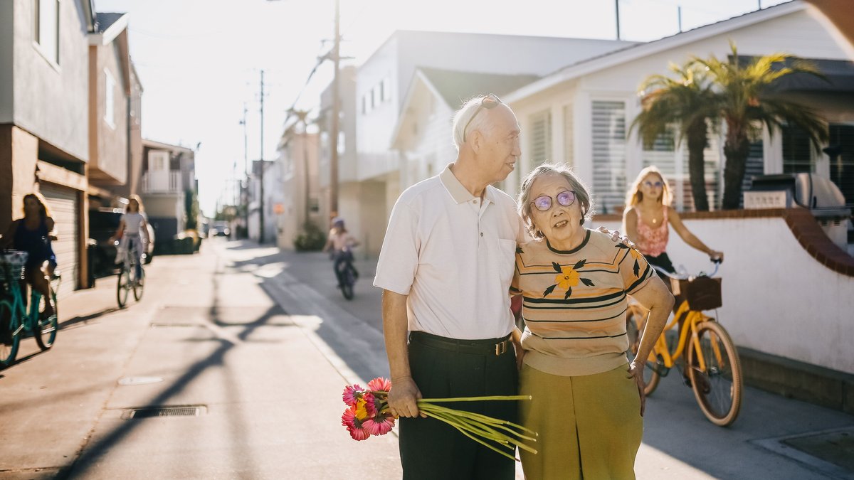 Elderly couple enjoying a walk with flowers in a sunny street setting, surrounded by cyclists.