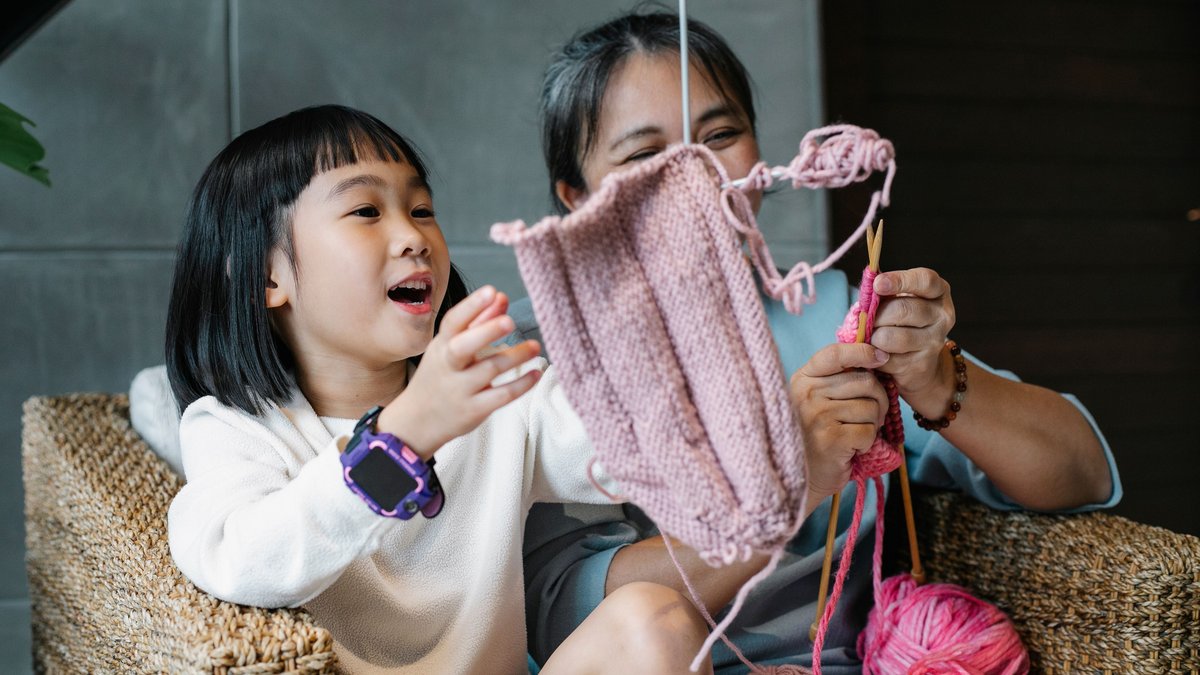 Charming Asian girl with laughing woman in armchair at home knitting together and having fun