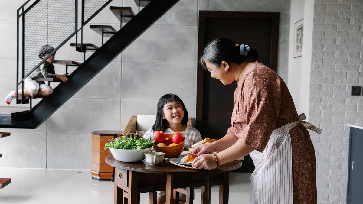 Side view of happy Asian grandmother serving delicious lunch with pasta and fresh vegetables for adorable little children while spending time together at home
