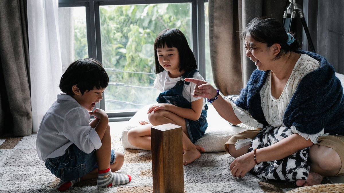 Cheerful aged ethnic woman sitting on carpet at home and having fun with adorable grandchildren while playing board game together