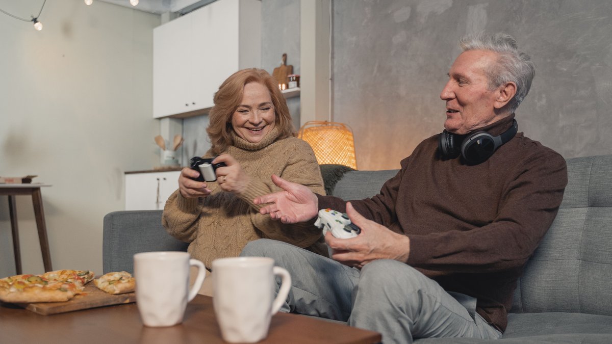 A joyful elderly couple playing video games with pizza and mugs nearby.