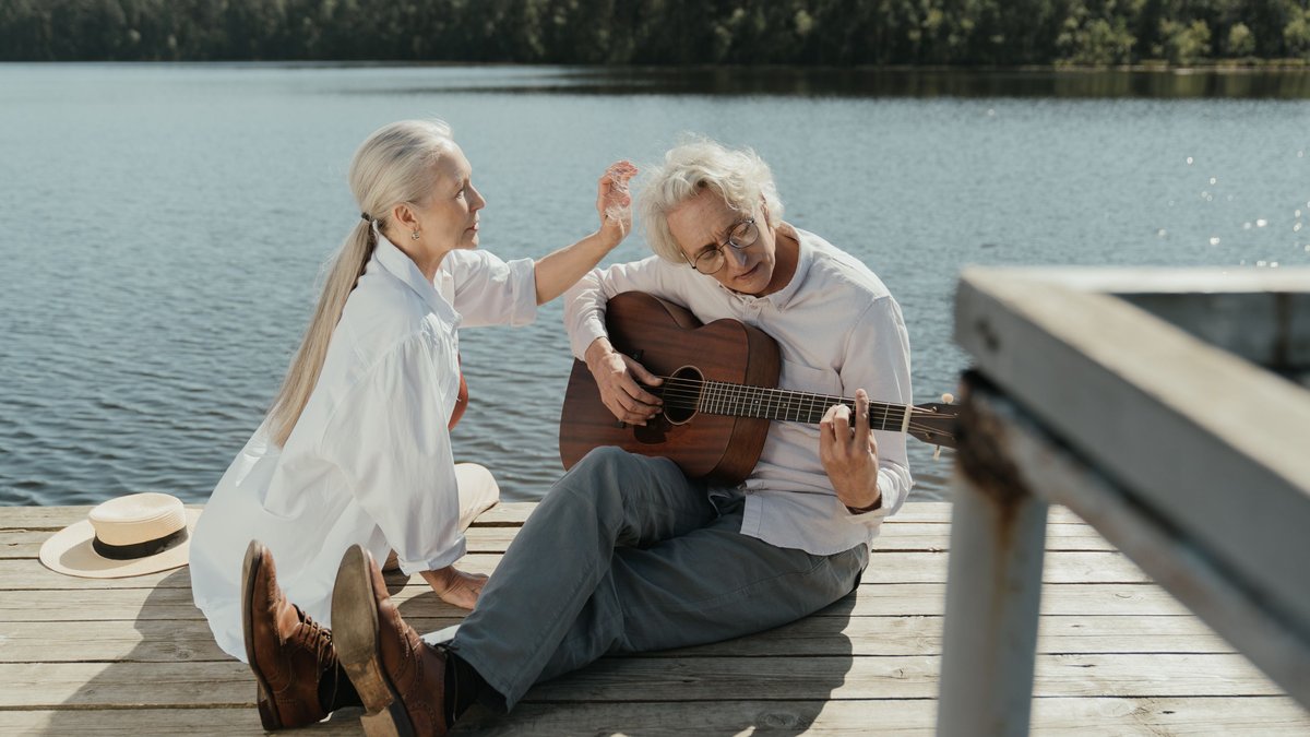 An elderly couple relaxes by the lake, playing guitar under the summer sun.