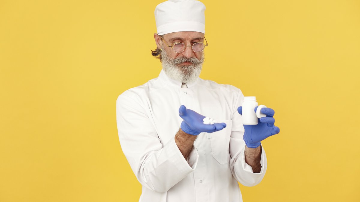 Senior pharmacist with beard and glasses holding medicine bottle and tablets on a yellow background.