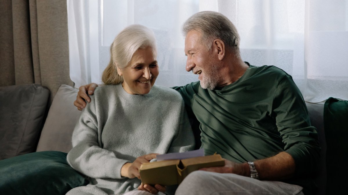 Senior couple embracing and reading a book on a sofa, sharing a joyful and intimate moment indoors.