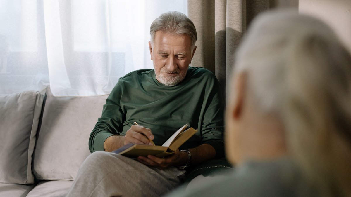 Senior man in a green sweater reading a book on a sofa, enjoying a peaceful indoor setting.