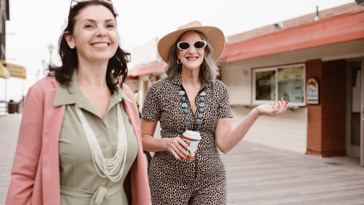 Two stylish senior women walking outside, smiling and enjoying coffee on a casual day out.