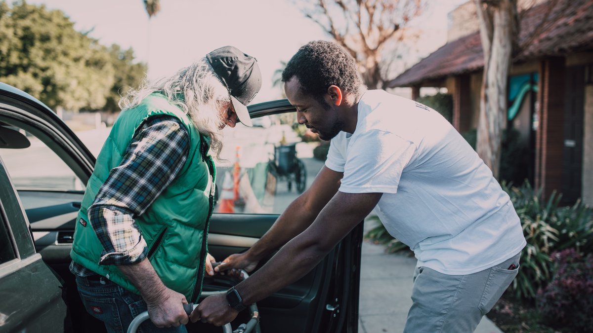 Young volunteer helping senior man with walker near a parked car, symbolizing care and support.