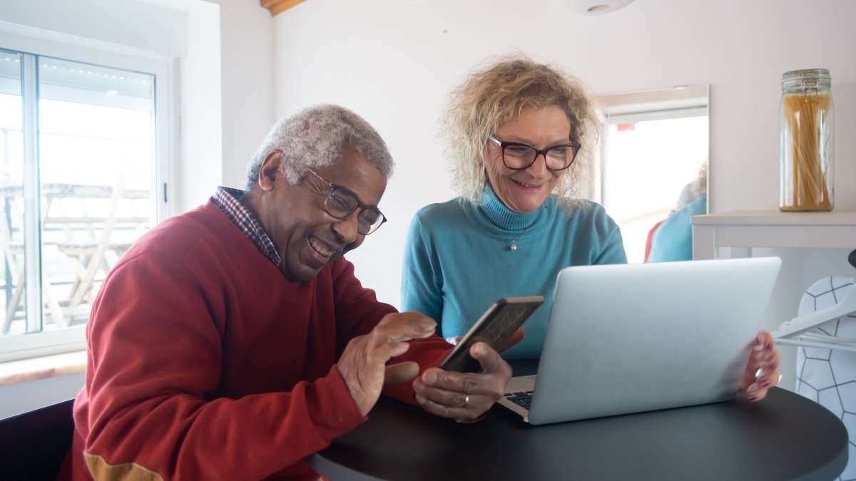 Smiling elderly couple using a smartphone and laptop indoors, enjoying online activities.