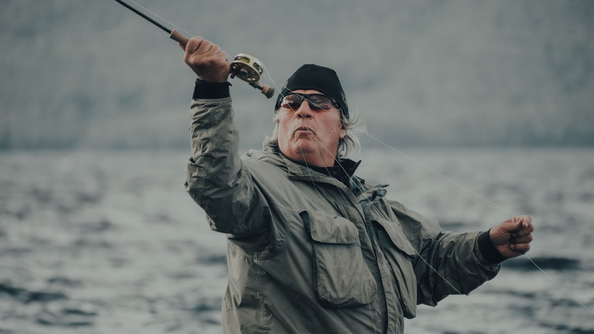 An elderly man in sunglasses enjoys fishing as a leisure activity in a tranquil lakeside environment.
