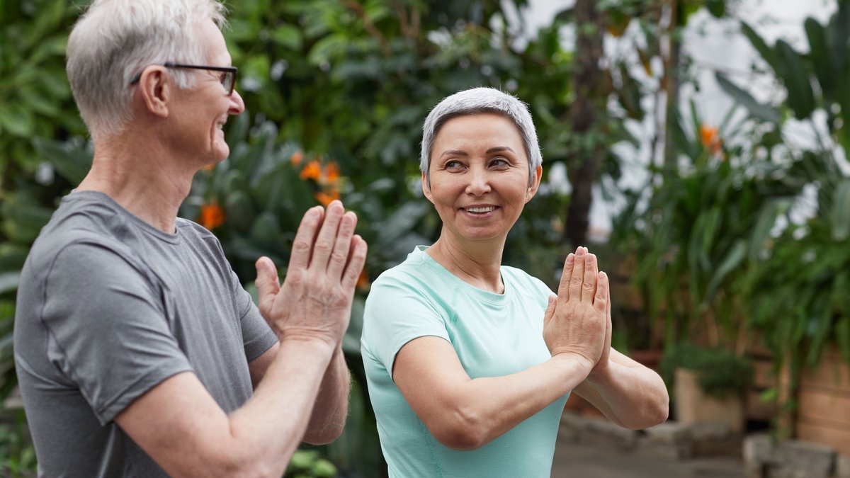 Happy senior couple practicing yoga in a lush greenhouse setting, embodying wellness and togetherness.