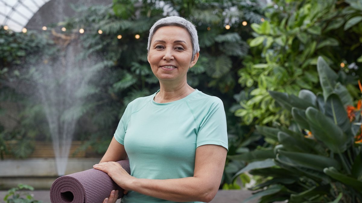 Elderly woman holding yoga mat and smiling in a lush greenhouse setting.