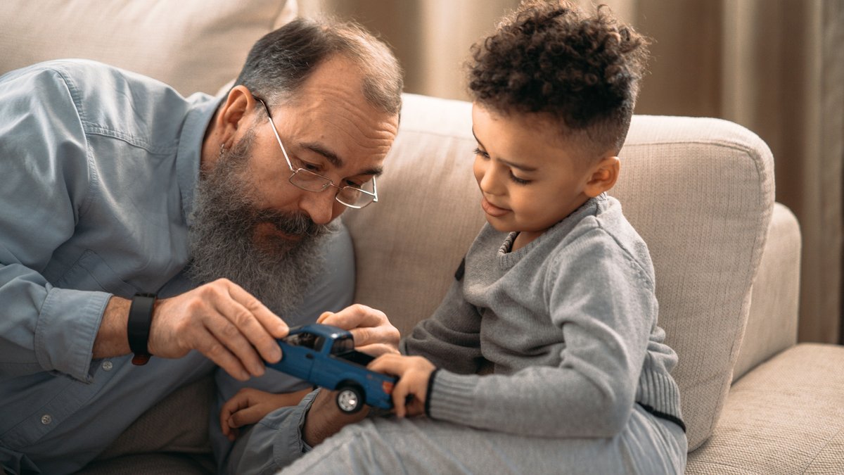 An elderly man and young boy bond while playing with a toy car indoors, showcasing family love.