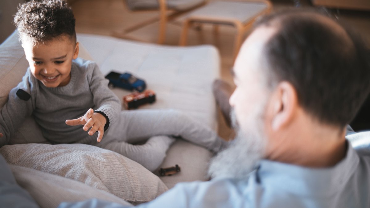 A cheerful grandfather and grandson sitting on a couch playing with toy cars in a living room setting.