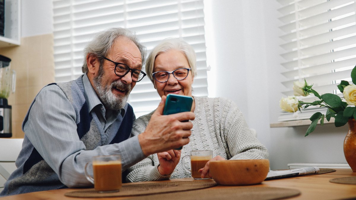 Happy senior couple sitting at a table, enjoying coffee and looking at a smartphone together in their cozy kitchen.