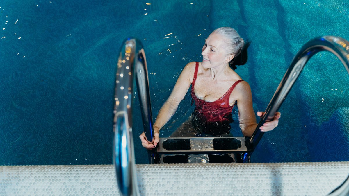 Elderly woman in a red swimsuit smiling while exiting a swimming pool.