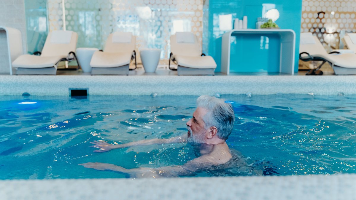 Older man enjoying a swim in a stylish indoor pool facility with relaxation chairs.