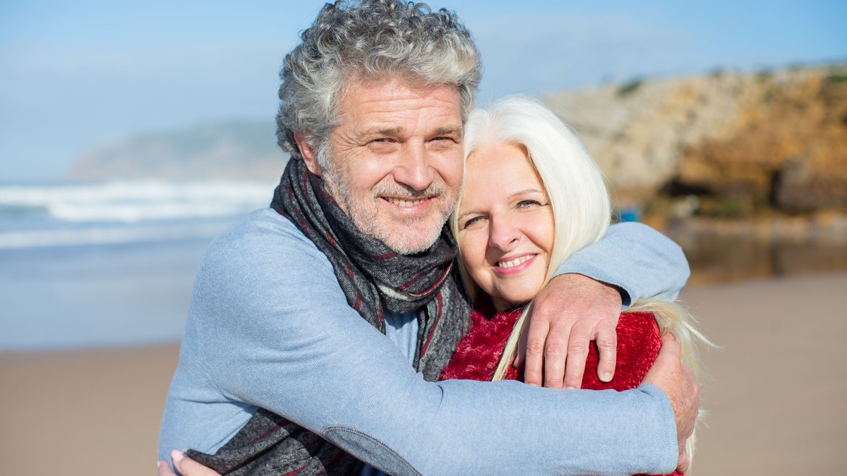 Joyful elderly couple hugging on a sunny beach in Portugal, enjoying a romantic day.