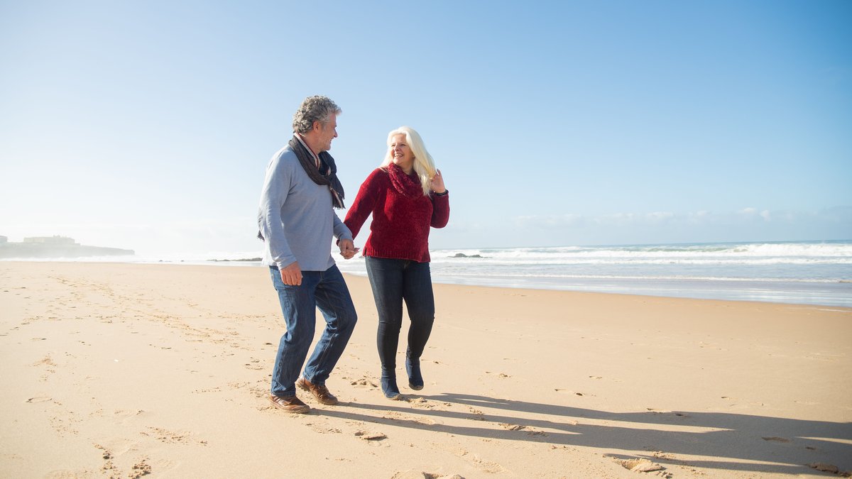 A happy senior couple holds hands while walking along a sunny beach in Portugal, enjoying a romantic moment.