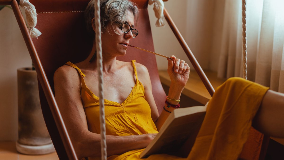 A senior woman in a hammock reading a book with a relaxed expression, enjoying leisure indoors.