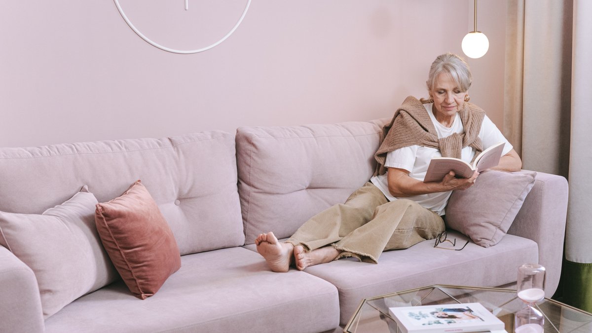 Senior woman relaxing and reading a book at home on a comfortable pink sofa.