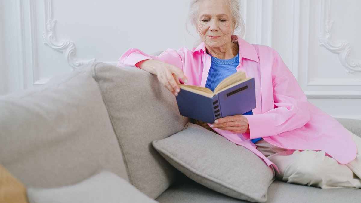 An elderly woman in a pink shirt enjoys relaxing on a couch while reading a book indoors.
