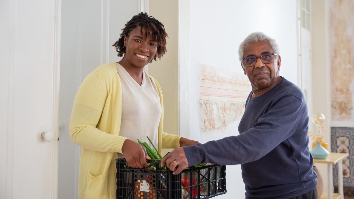 Smiling woman assists a senior man with food delivery indoors, promoting community support and care.