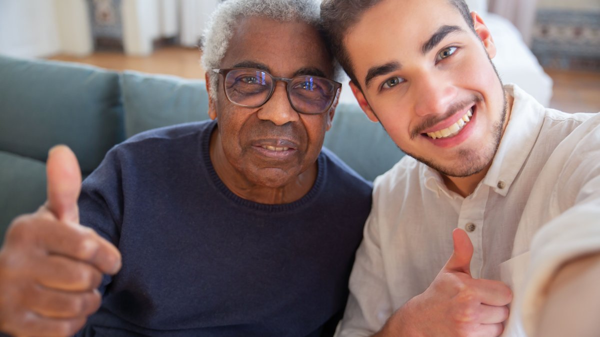 An elderly man and a young man smiling with thumbs up at home, representing friendship and care.