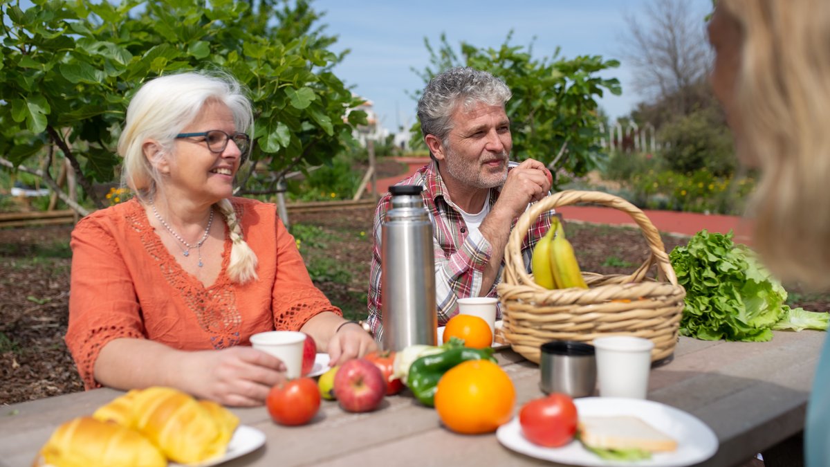 Senior couple enjoys a cheerful picnic with fresh fruits and vegetables in a lush garden setting.