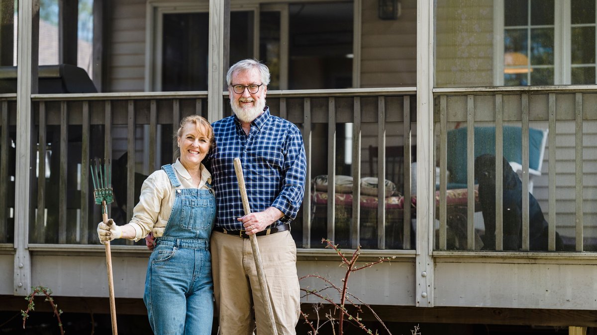 Elderly couple joyfully standing outside their home with gardening tools, symbolizing happiness and love.
